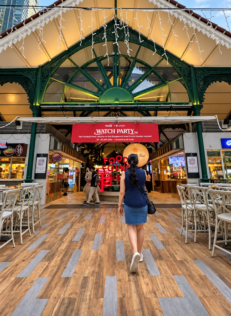 A woman walks towards a vibrant entrance of a market with a 'Watch Party' banner, surrounded by food stalls and festive decorations.